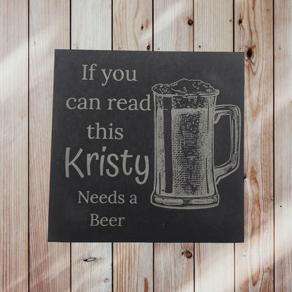 personalised slate drinks coaster with 'If you can read this, Kristy Needs a Beer' text on a light wooden background