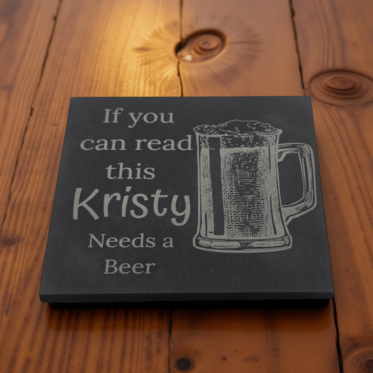 Slate coaster with a beer glass engraving and funny quote text on a wooden table.