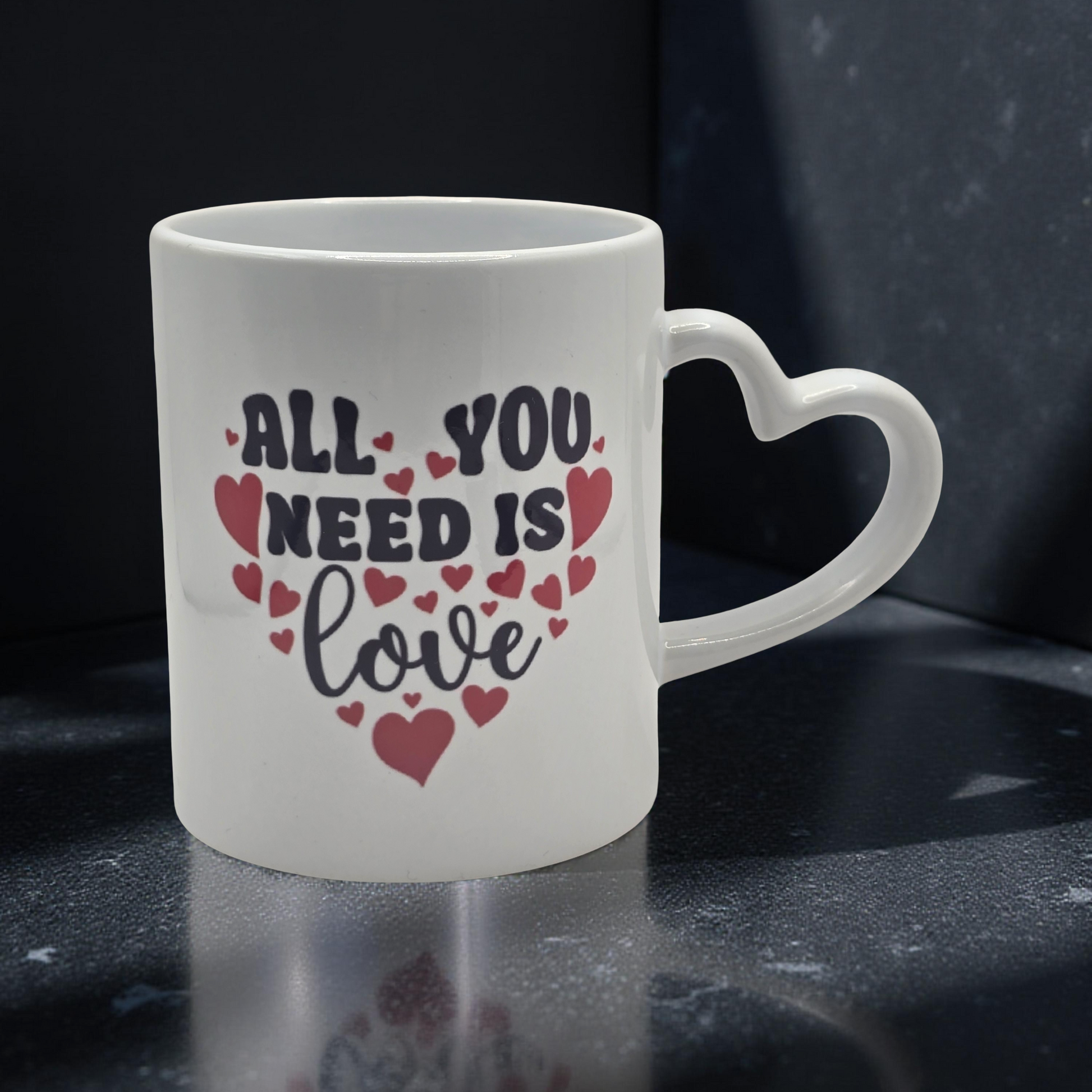 White mug with 'All You Need Is Love' text and heart designs on a dark surface