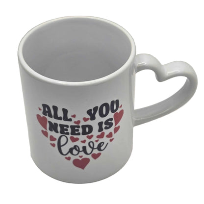 White mug with heart-shaped handle and 'All You Need Is Love' text on a white background