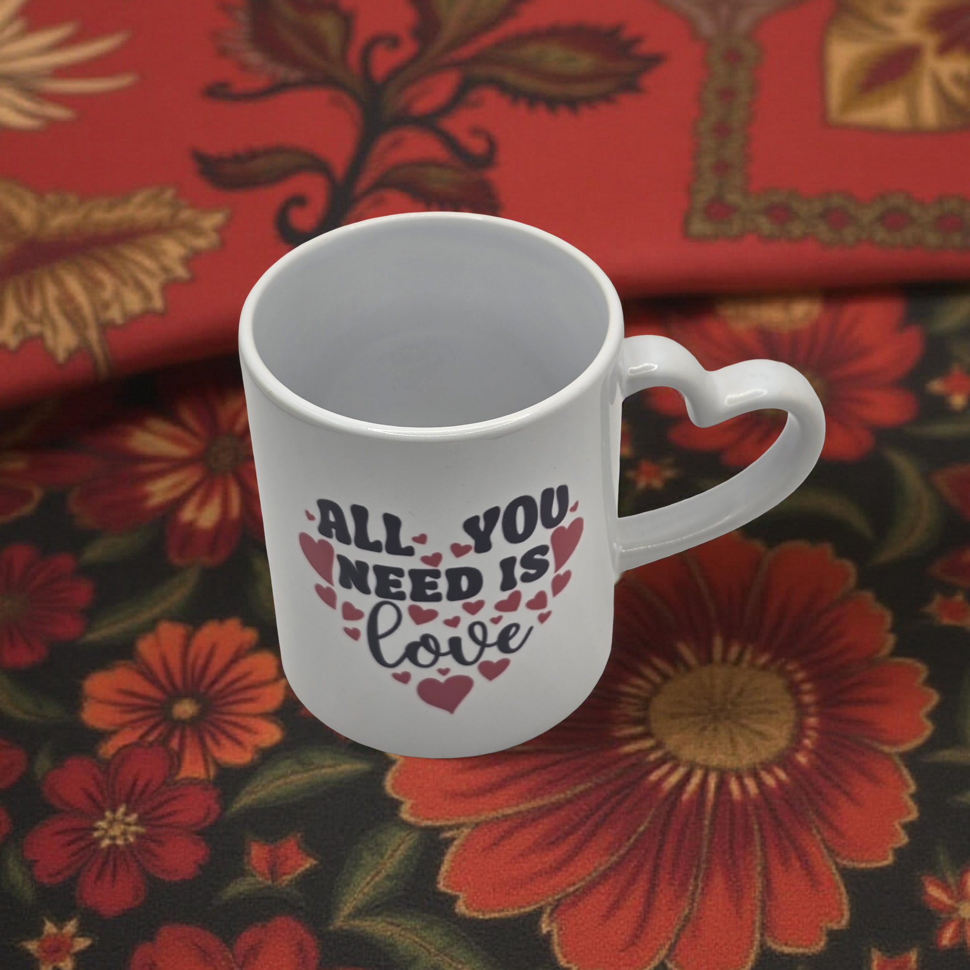White mug with heart handle featuring 'All You Need Is Love' text on a floral fabric background
