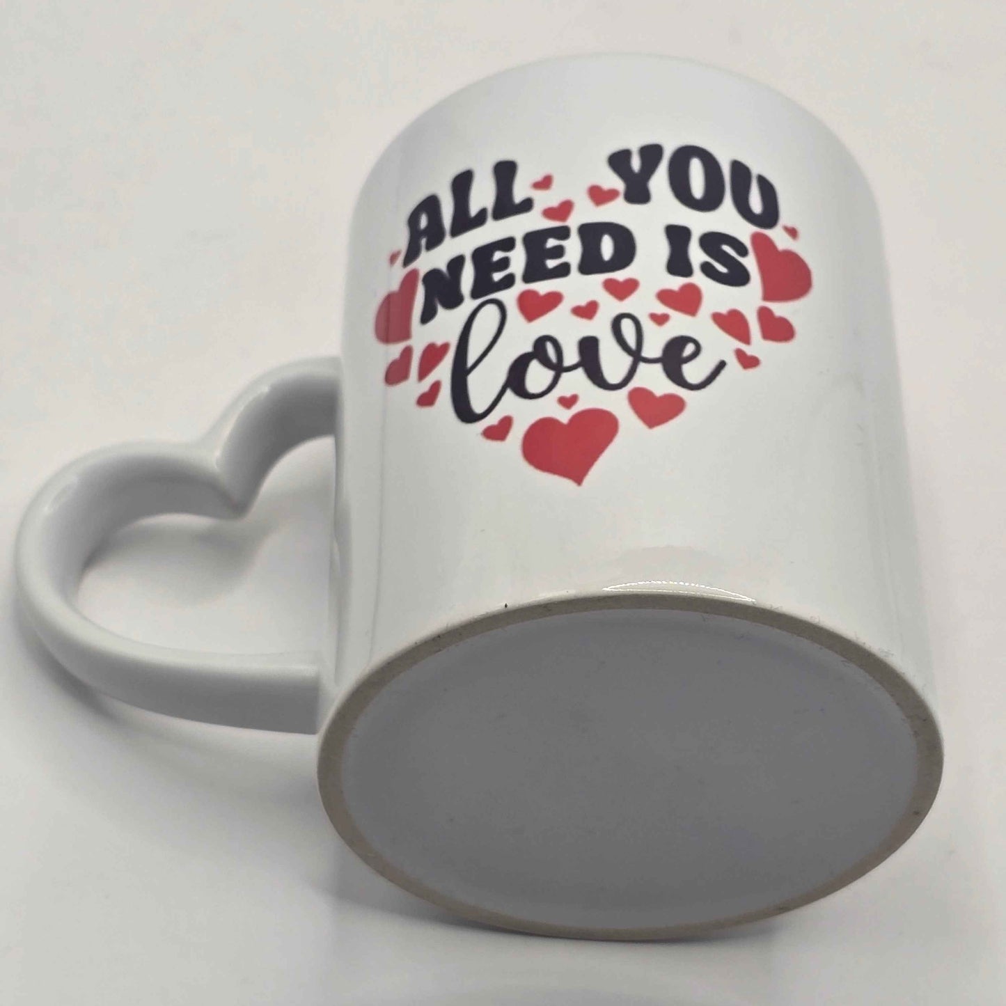 White mug with 'All You Need Is Love' text and heart designs on a white background showing the underside of the mug