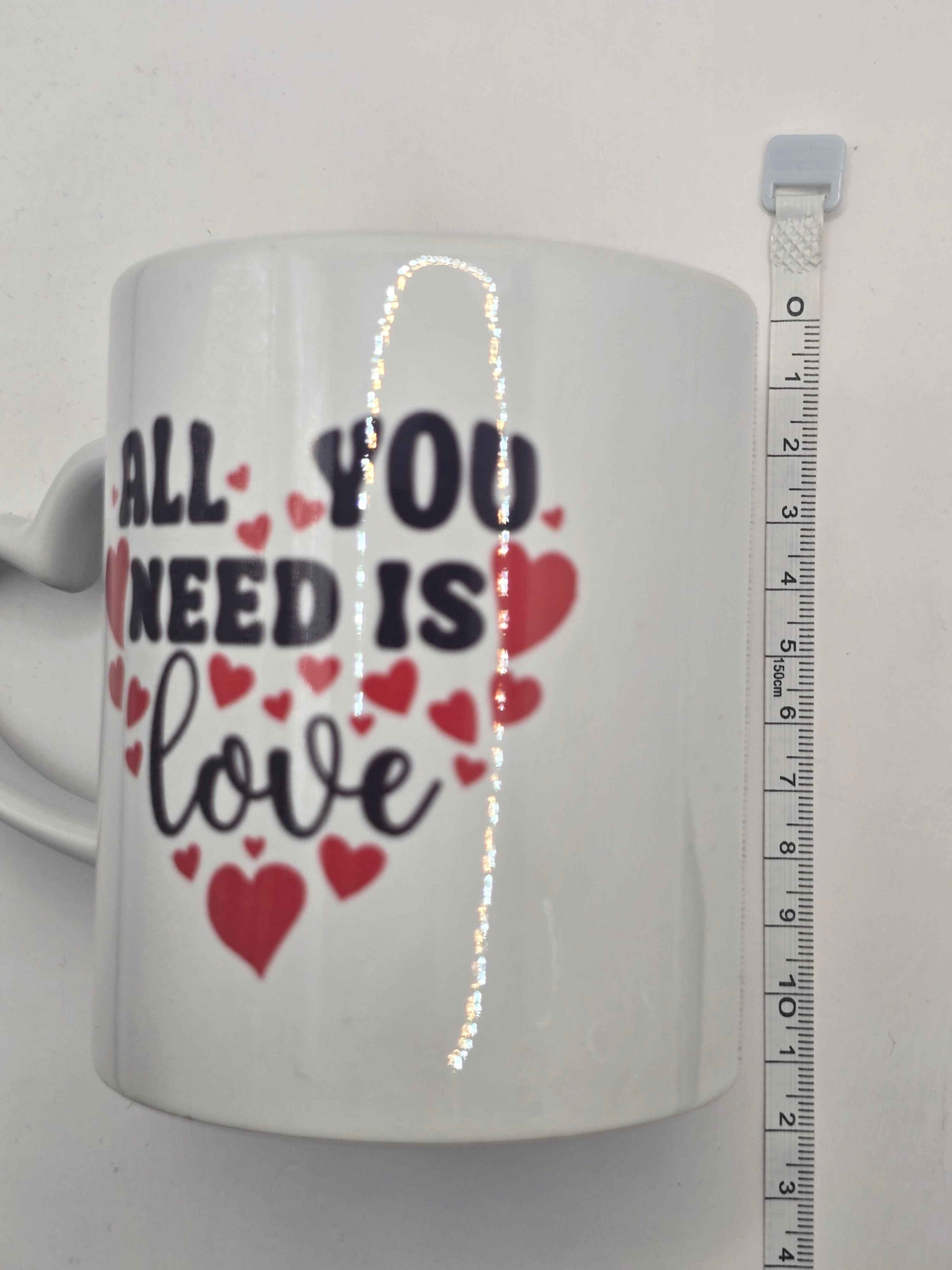 White mug with 'All you need is love' text and hearts, next to a ruler for size reference.