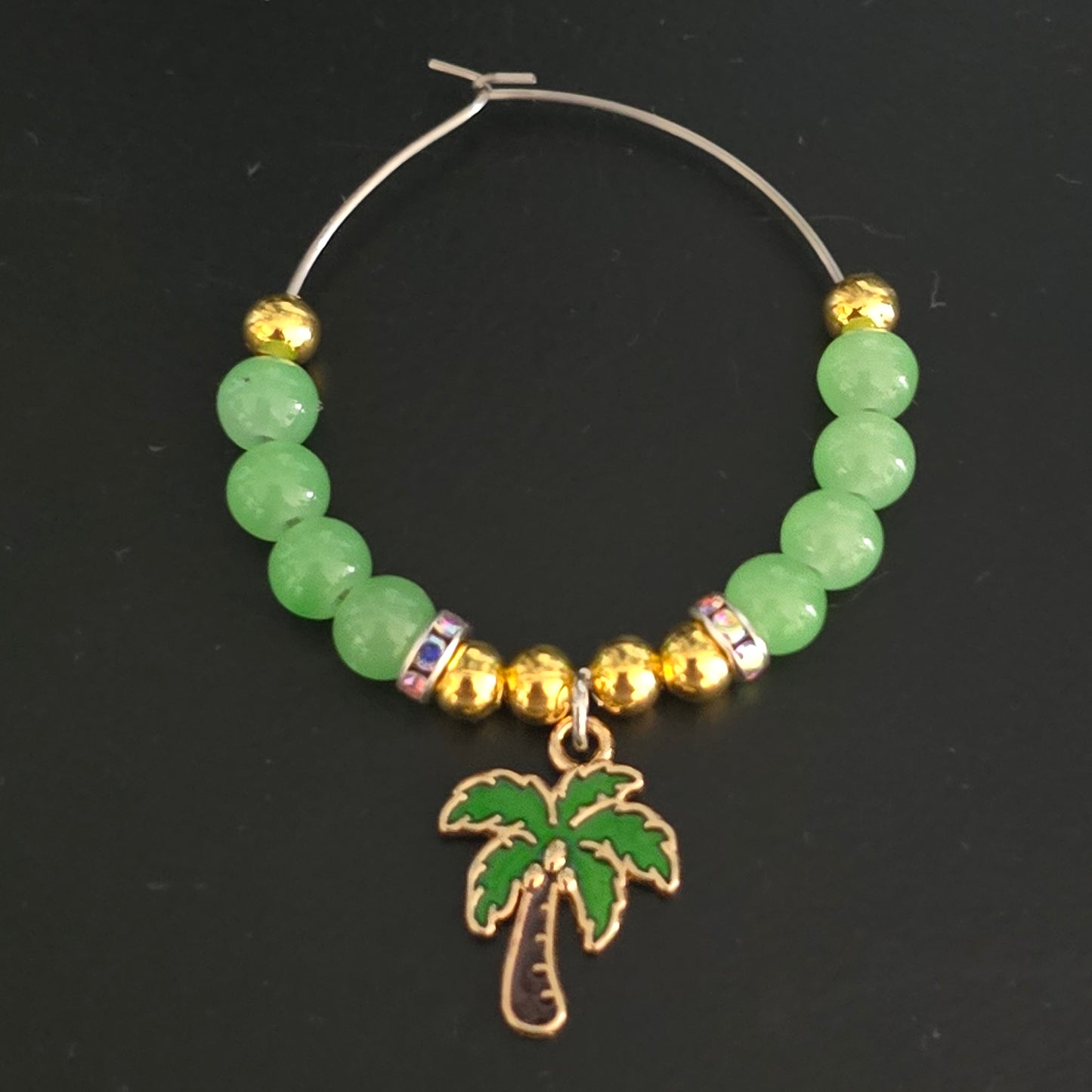 top down shot of a tropical themed wine glass charm with a palm tree charm, green and gold beads