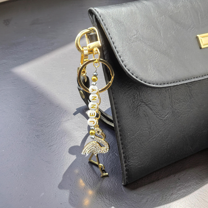 Gold flamingo keychain with rhinestones and gold and white lettered beads attached to a black handbag