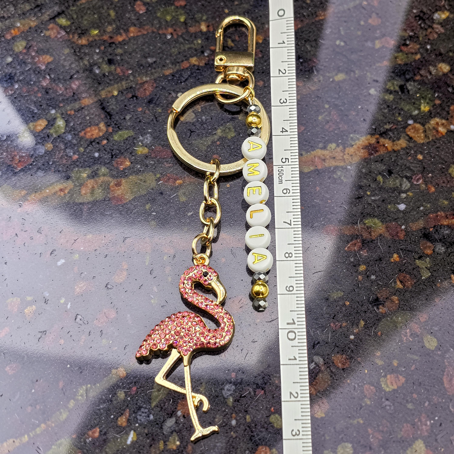 Flamingo keychain with 'AMELIA' beads next to a ruler on a textured surface