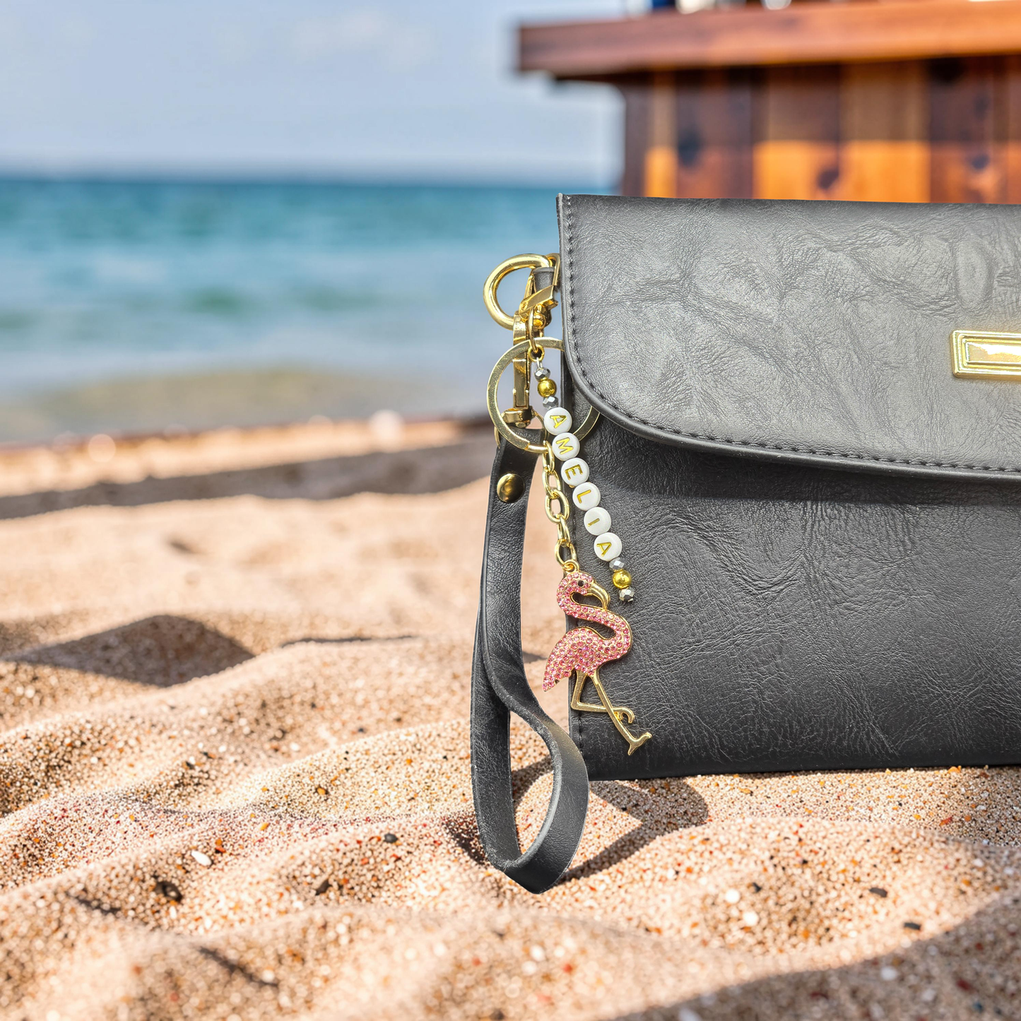 Black handbag with a personalised flamingo keychain on a sandy beach with ocean in the background