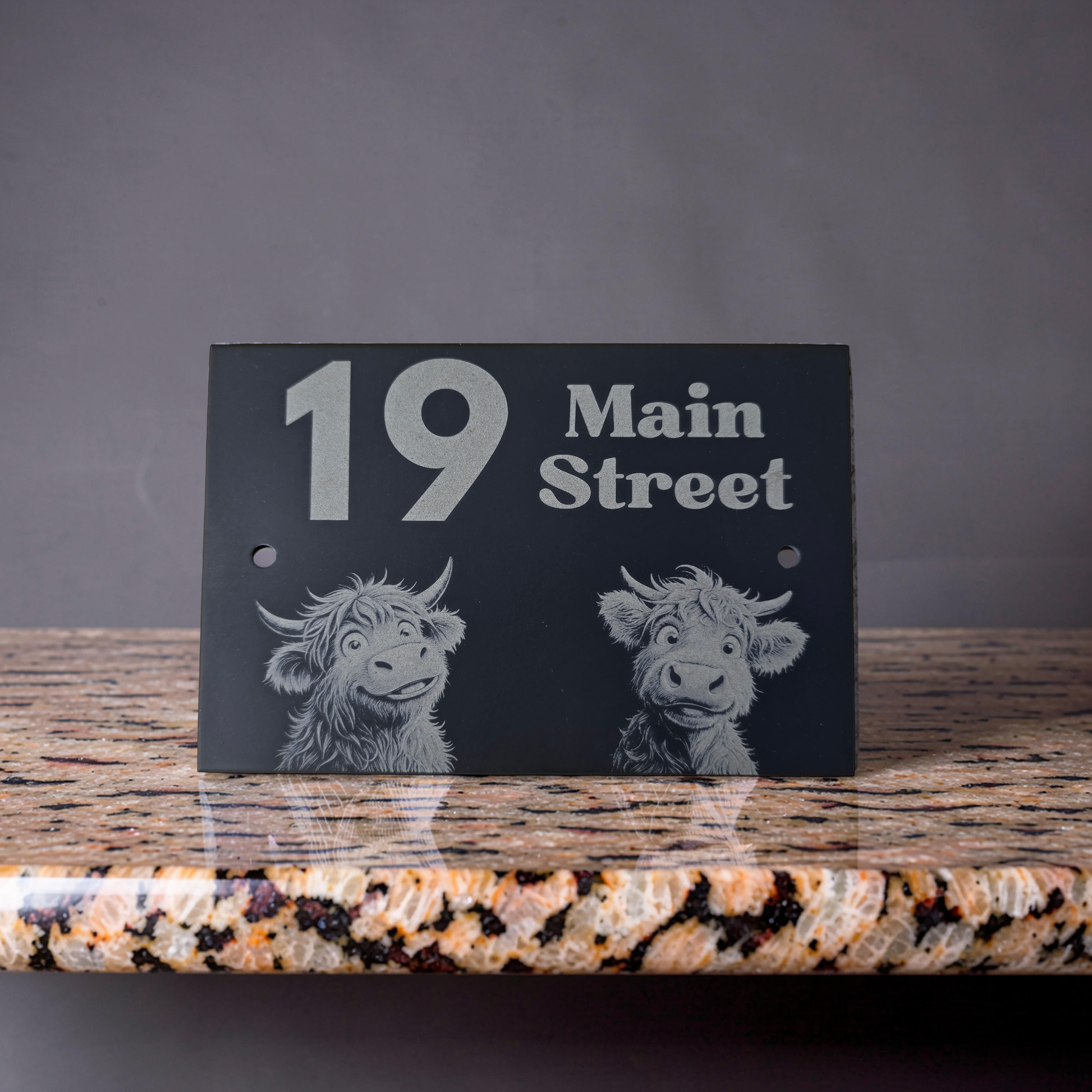 Black slate house address sign with white text and highland cow illustrations on a granite surface