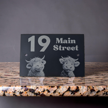 Black slate house address sign with white text and highland cow illustrations on a granite surface