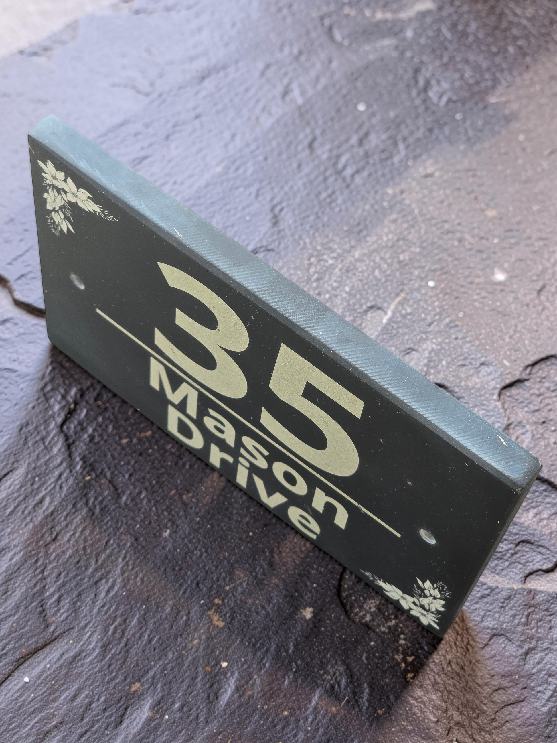 A rectangular black house number sign reading '35 Mason Drive' with small white floral corner accents, photographed on a dark textured stone surface.