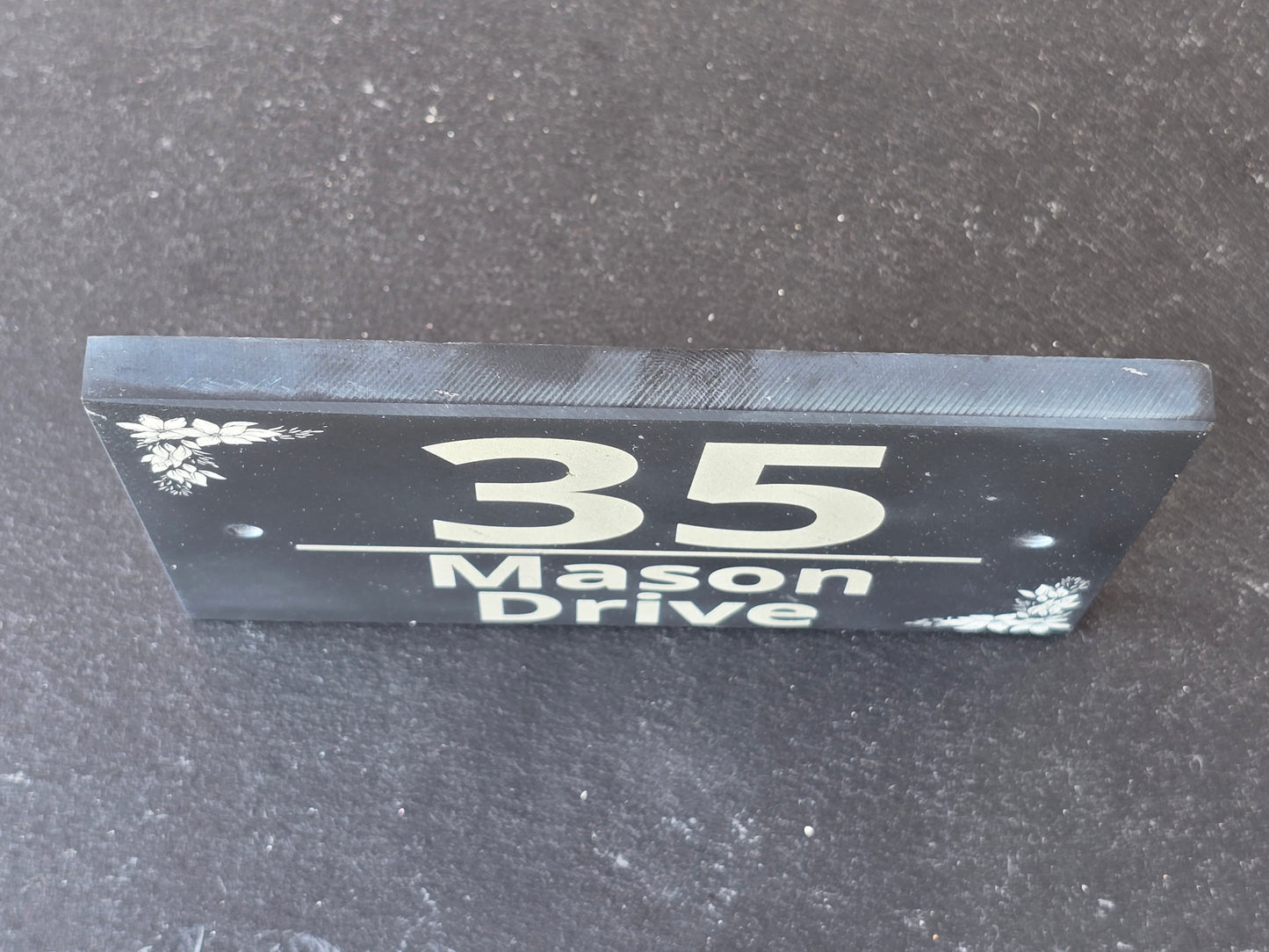A black rectangular house number sign with white lettering reading 'Mason Drive 35' and small white floral corner details, photographed standing upright on a dark textured surface.