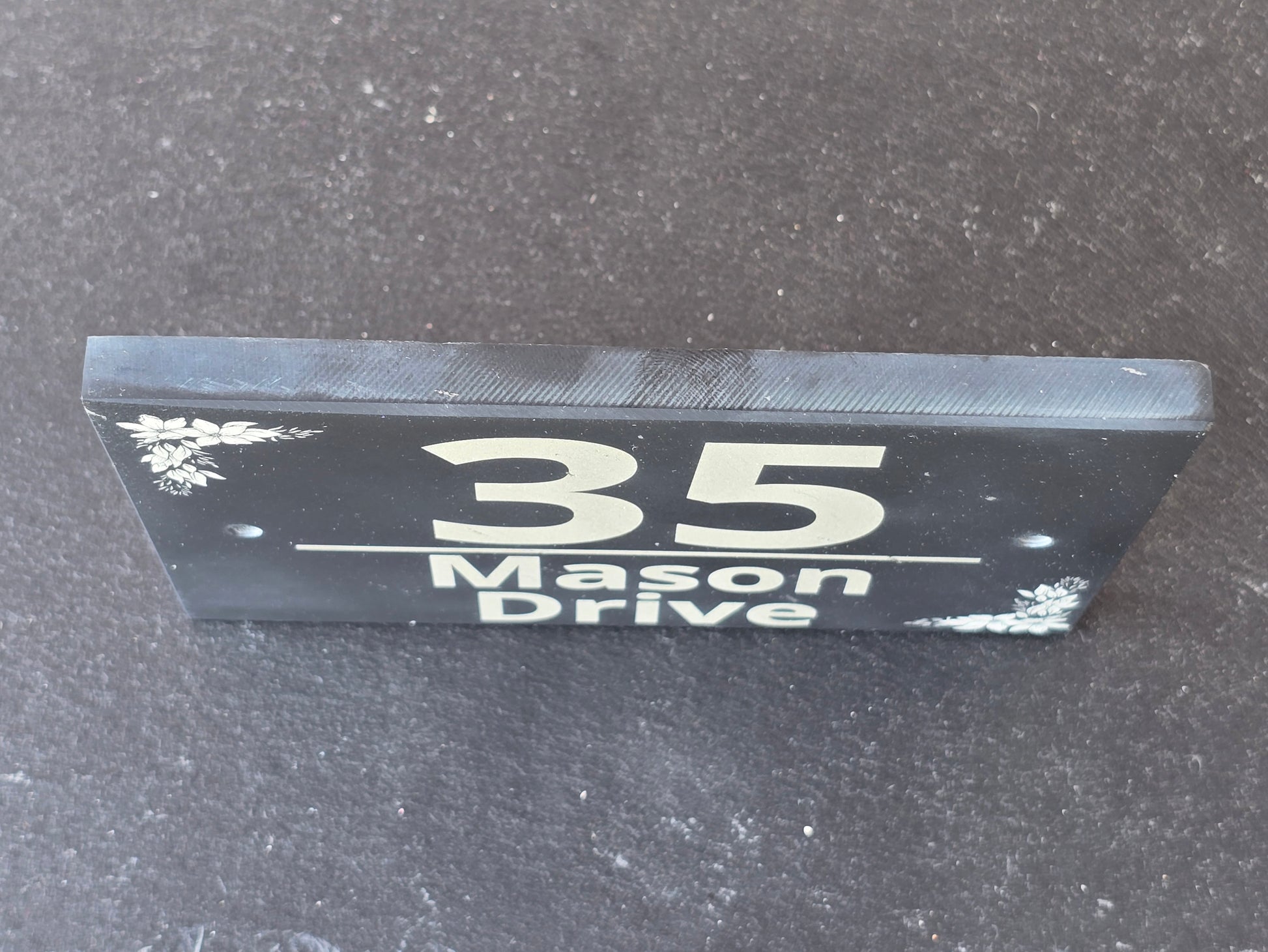A black rectangular house number sign with white lettering reading 'Mason Drive 35' and small white floral corner details, photographed standing upright on a dark textured surface.