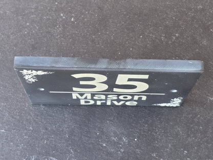 A black rectangular house number sign with white lettering reading 'Mason Drive 35' and small white floral corner details, photographed standing upright on a dark textured surface.