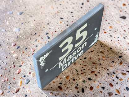A rectangular dark gray wooden house number sign displaying '35 Mason Drive' in light gray lettering, shown standing upright on a speckled terrazzo floor.