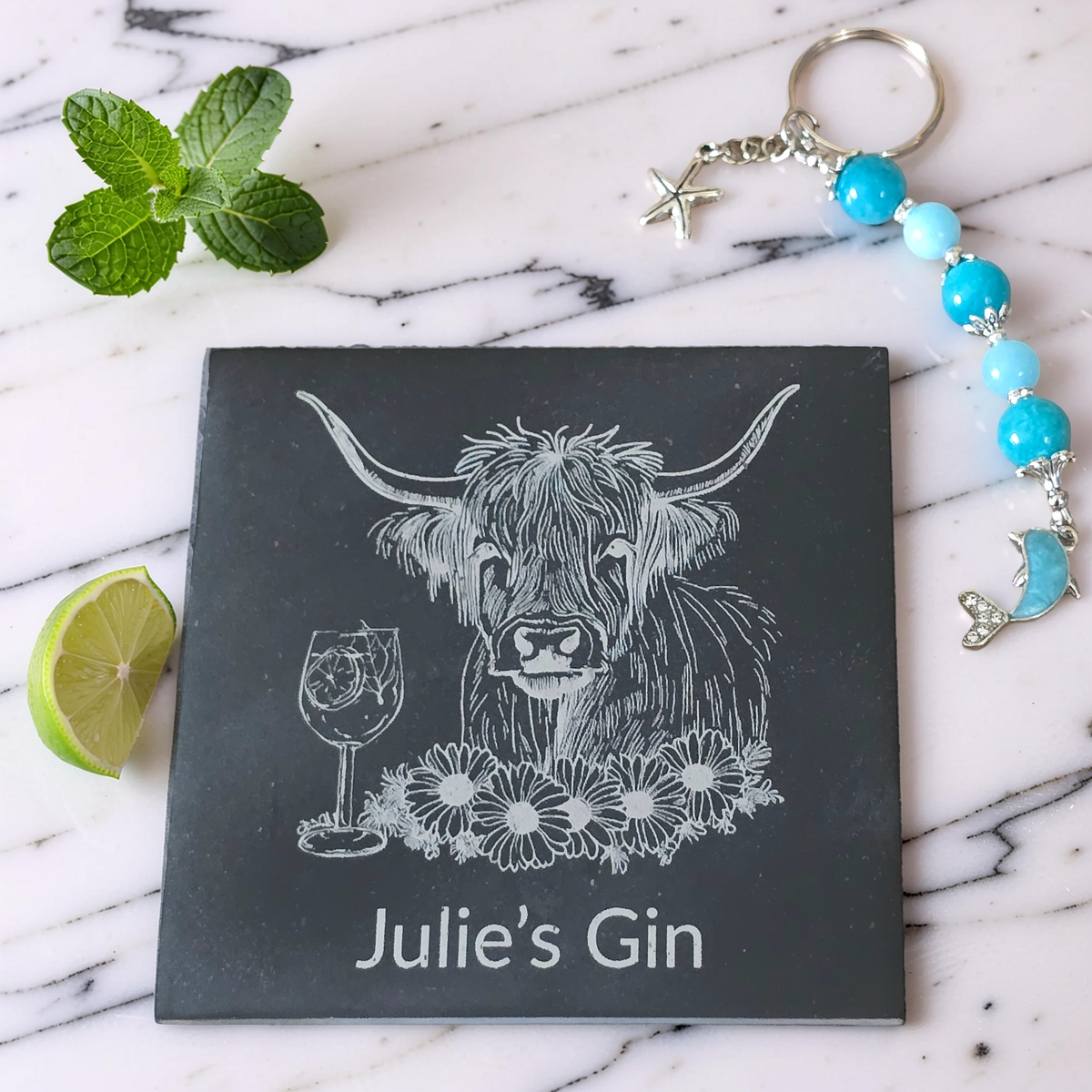 A square dark slate coaster with a white engraved illustration of a Highland cow, a wine glass, flowers, and the custom text 'Julie's Gin', styled on a white marble surface with mint leaves, lime, and a beaded keychain with turquoise beads.