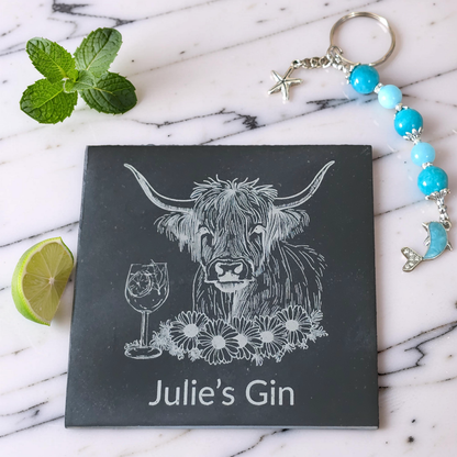 A square dark slate coaster with a white engraved illustration of a Highland cow, a wine glass, flowers, and the custom text 'Julie's Gin', styled on a white marble surface with mint leaves, lime, and a beaded keychain with turquoise beads.