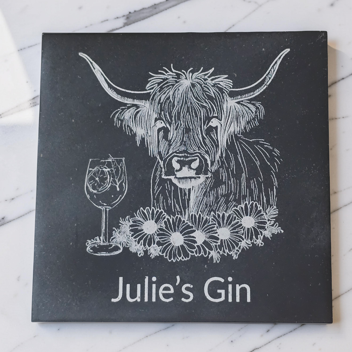 A square black slate coaster featuring a white line-art illustration of a Highland cow, a wine glass, flowers, and the text 'Julie's Gin'. The item is photographed flat on a white marble surface.