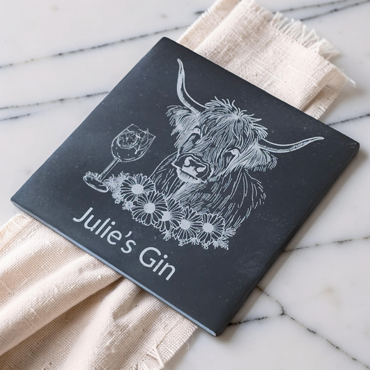 A personalised square black slate coaster or tile with a white engraved illustration of a Highland cow, a wine glass, flowers, and the text 'Julie's Gin', styled on a cream linen napkin over a white marble surface.