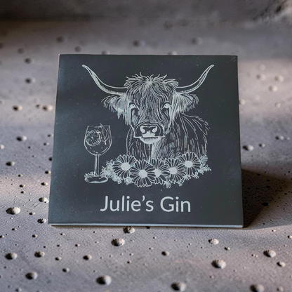 A square dark slate coaster featuring a white engraved illustration of a Highland cow, a wine glass, flowers, and the personalised text 'Julie's Gin'.