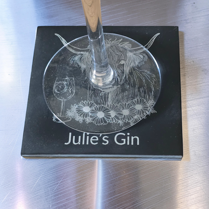 A square black coaster engraved with a Highland cow, flowers, a wine glass, and the text 'Julie's Gin', with a wine glass resting on top, set on a brushed stainless steel surface.