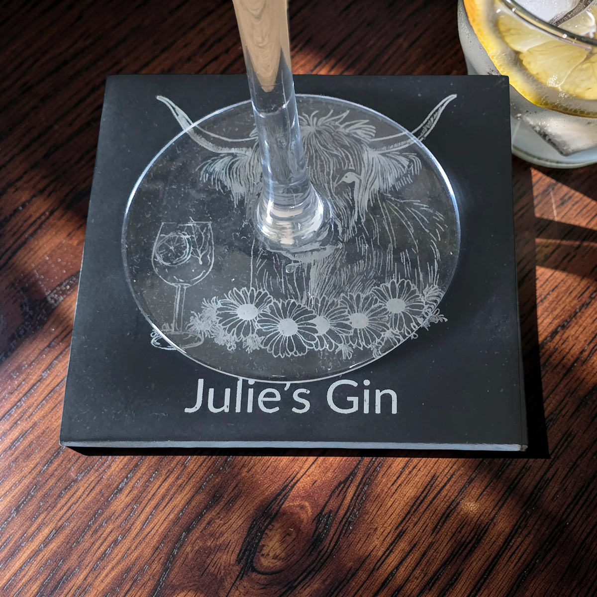 A square black slate coaster engraved with a Highland cow, flowers, a wine glass, and the text 'Julie's Gin'. place on a dark wooden table
