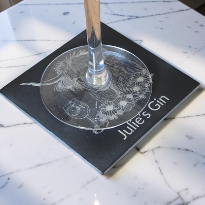 A Highland cow design personalised coaster with custom text sits on a white marble surface. the bottom of a wine glass sits on the coaster