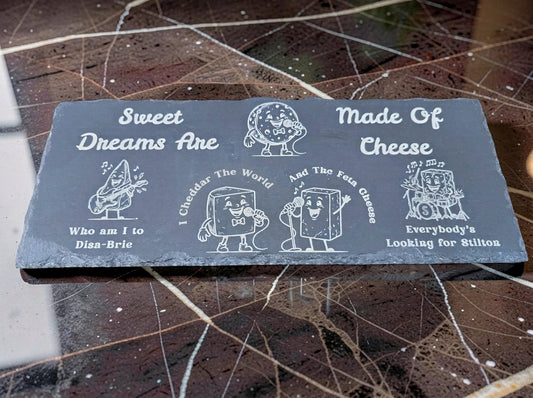 Rectangular slate board with chipped edges, engraved with cartoon cheese musicians and pun song lyrics.