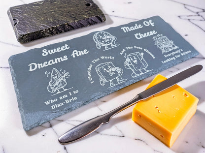 Engraved slate board with funny cheese puns, shown beside a cheese knife and cheddar block on a marble counter.