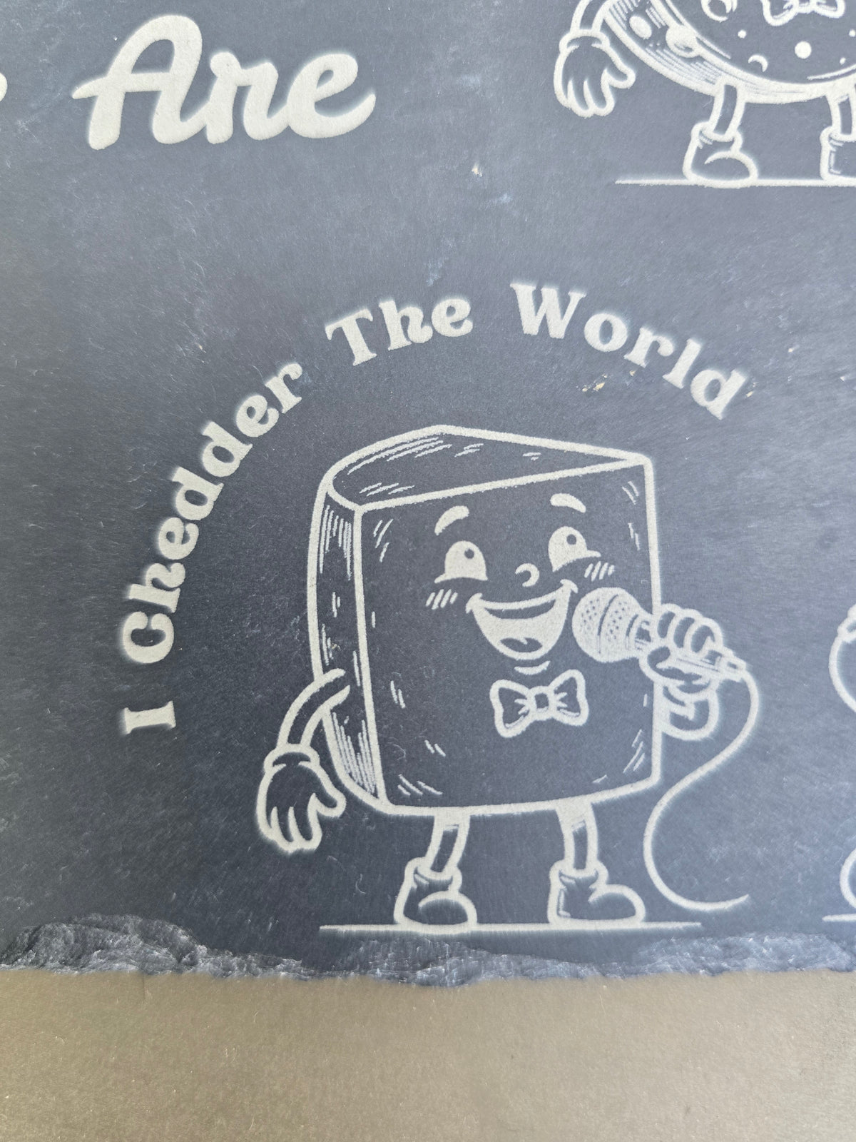 Engraved slate board detail of a cartoon cheese singing into a microphone under the words I Chedder The World.