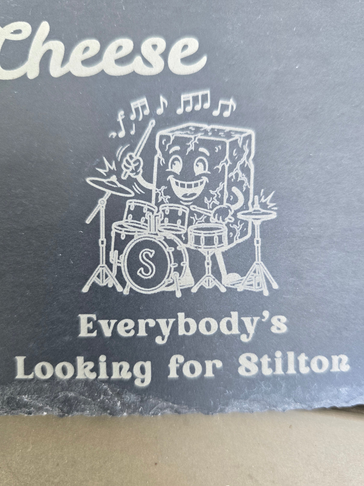Engraved slate detail of a cartoon cheese playing drums above the phrase Everybody's Looking for Stilton.