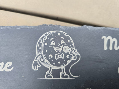 Engraved cartoon cheese wheel in a bowtie singing into a microphone on a dark slate board.