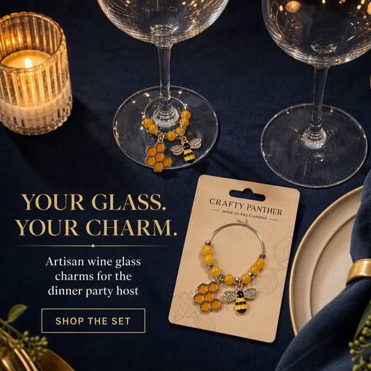 Two beaded wine glass charms with yellow honeycomb and bumblebee details are displayed on a dark navy tablecloth with wine glasses, plates, and a lit candle. One charm is shown on a card labeled 'Crafty Panther Mini Glass Charms' and the other is draped over a glass base.