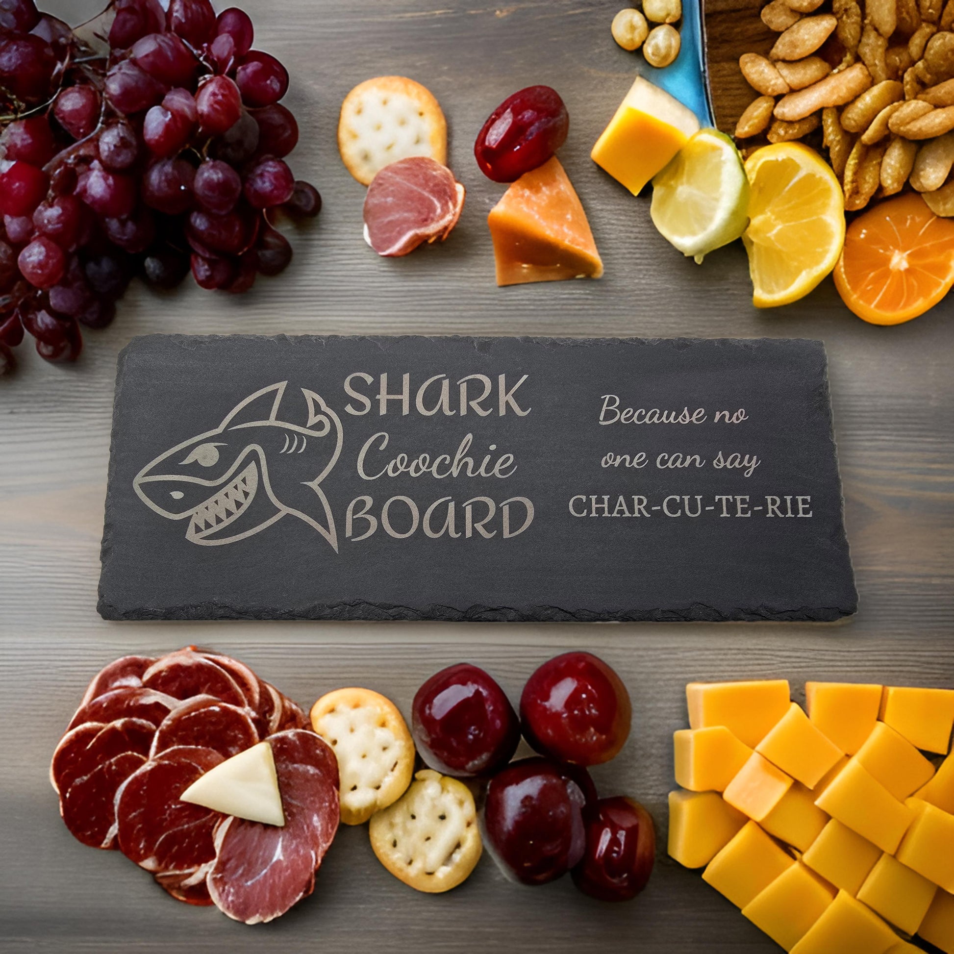 A slate charcuterie board with 'Shark Coochie Board' text and a shark illustration, surrounded by an assortment of snacks such as fruits, cheeses, and crackers.
