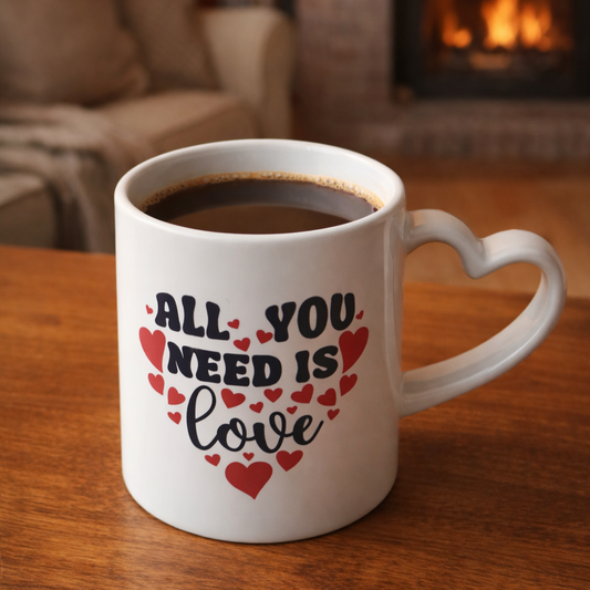 White mug with 'All you need is love' text on a wooden table in a cozy room. the mug has a heart shaped handle