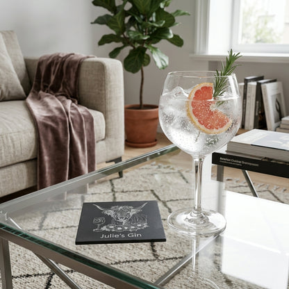 A glass of gin with a grapefruit slice and rosemary garnish sits on a glass coffee table beside a black square coaster printed with personalised text 'Julie's Gin' and a highland cow image . The scene is set in a modern living room with a sofa, plant, and books in the background.