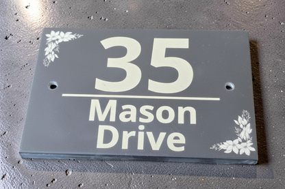 A rectangular black house number plaque with white lettering reading '35 Mason Drive' and small white floral corner accents, photographed on a textured gray surface.