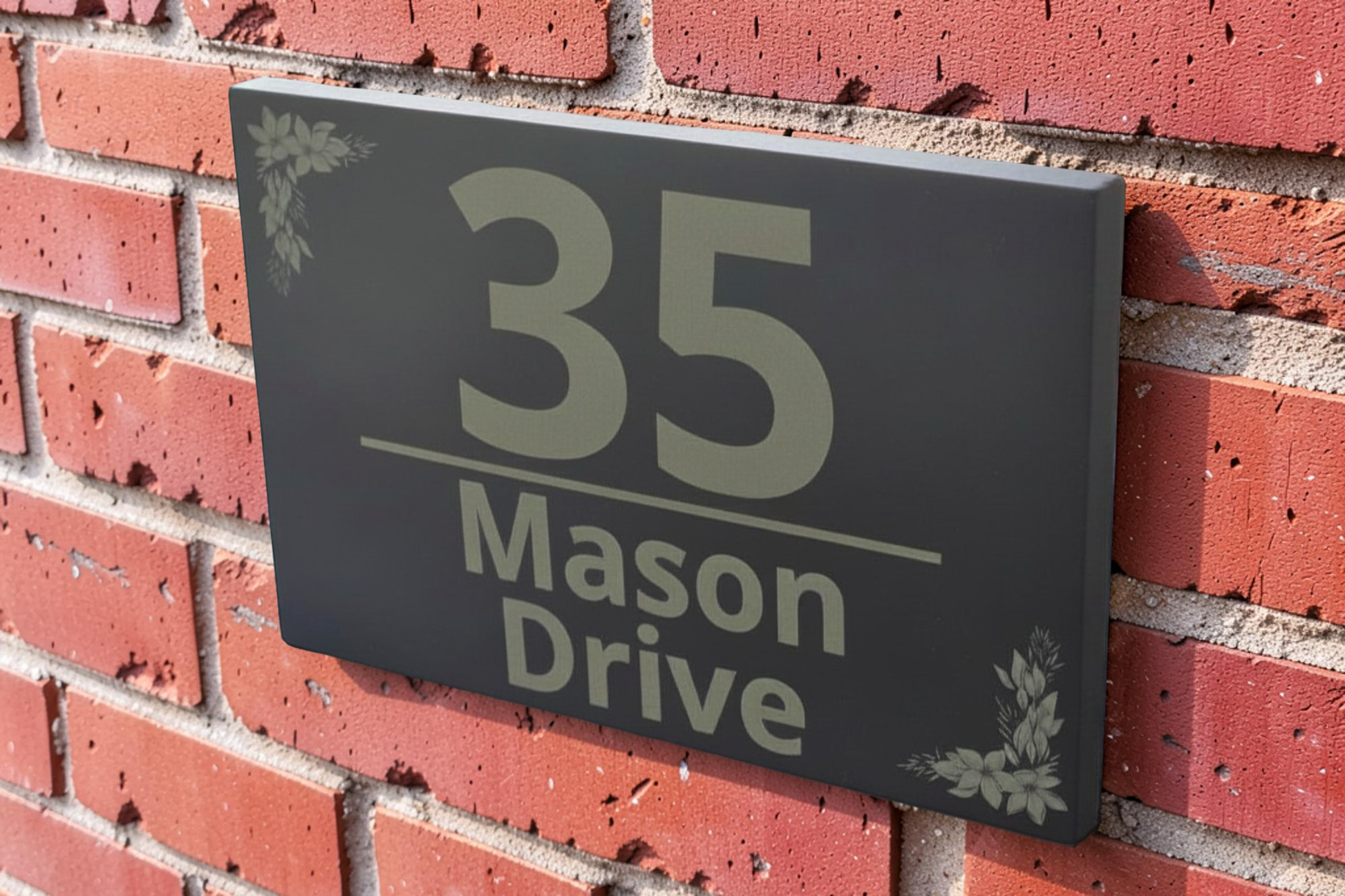 A rectangular black house number plaque mounted on a red brick wall, displaying '35 Mason Drive' in light gray lettering with small leaf corner accents.