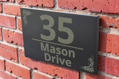 A rectangular black house number plaque mounted on a red brick wall, displaying '35 Mason Drive' in light gray lettering with small leaf corner accents.