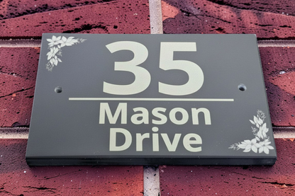 A rectangular black house number sign reading '35 Mason Drive' with small white floral corner accents, mounted on a red brick wall.