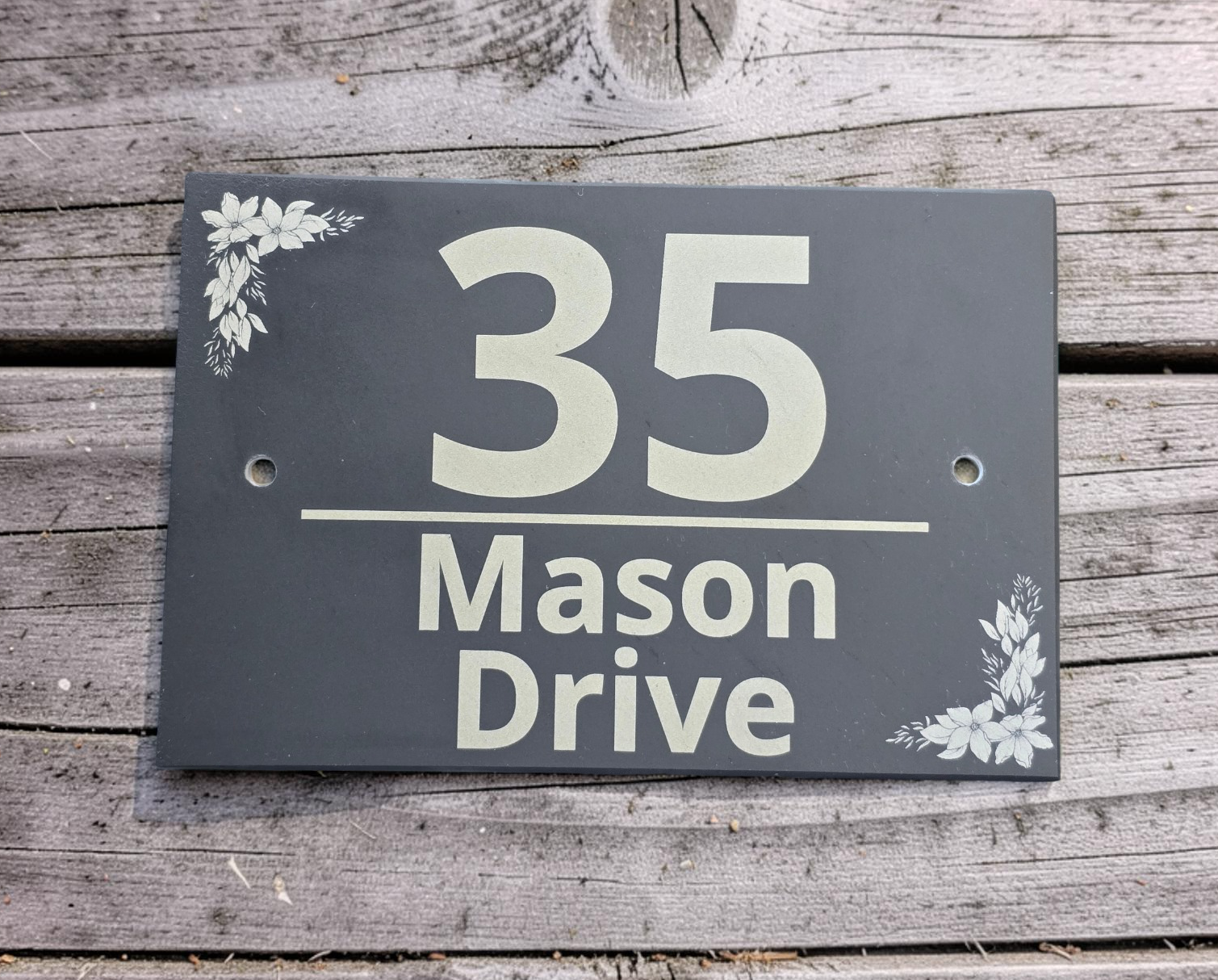 A rectangular dark gray house number sign reading '35 Mason Drive' with small white floral corner accents, mounted on weathered wood siding.