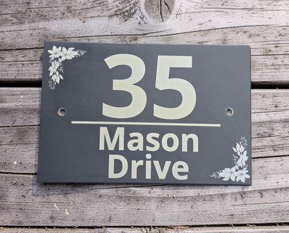 A rectangular dark gray house number sign reading '35 Mason Drive' with small white floral corner accents, mounted on weathered wood siding.