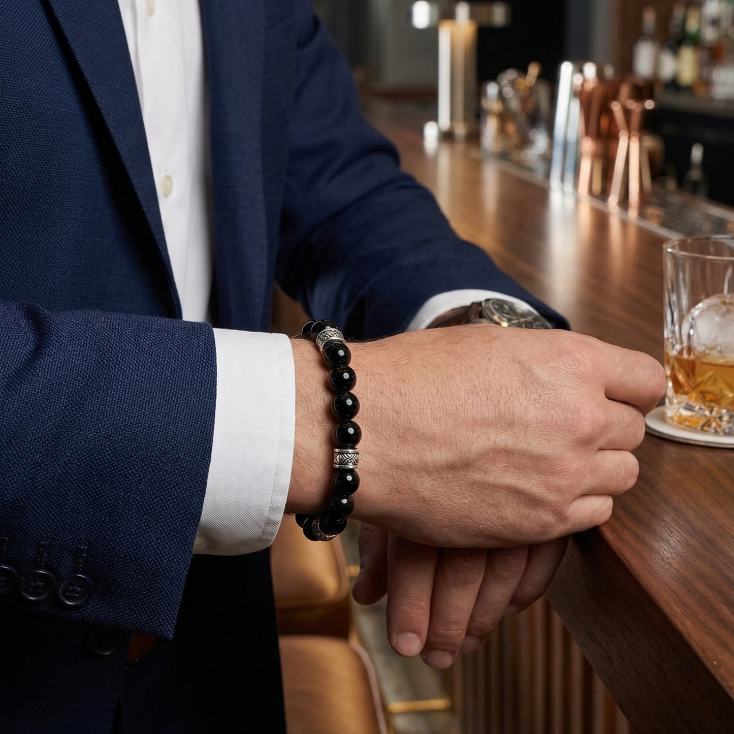 Person in a suit with a black gloss beaded bracelet holding a glass of whiskey at a bar.