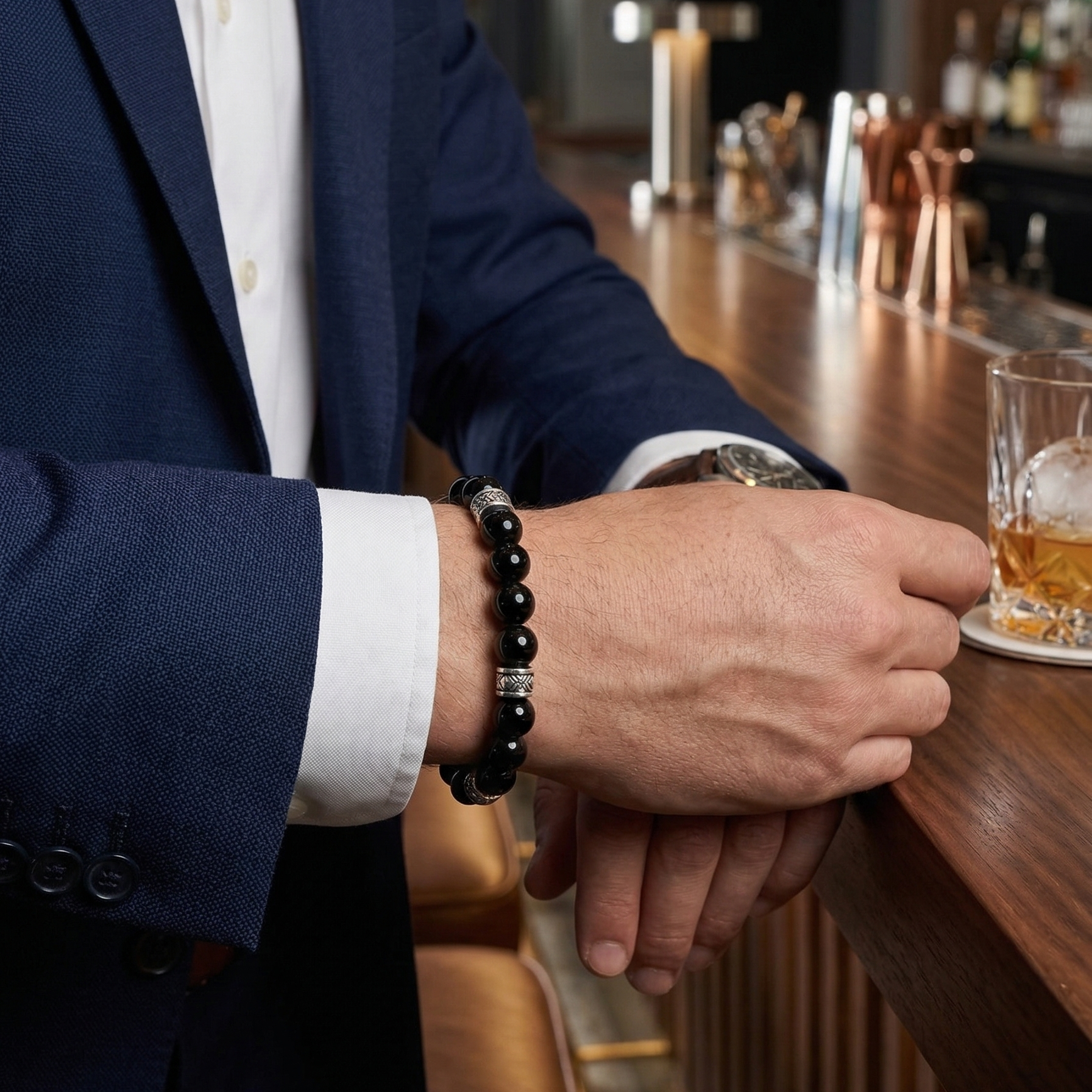 Person in a suit with a black gloss beaded bracelet holding a glass of whiskey at a bar.