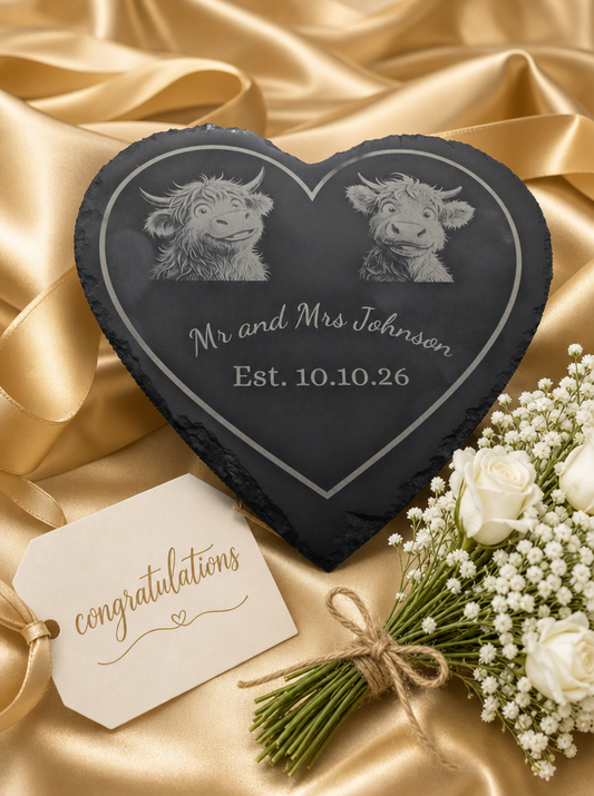 A heart-shaped black slate plaque engraved with two Highland cow portraits and the text 'Mr and Mrs Johnson" and a date