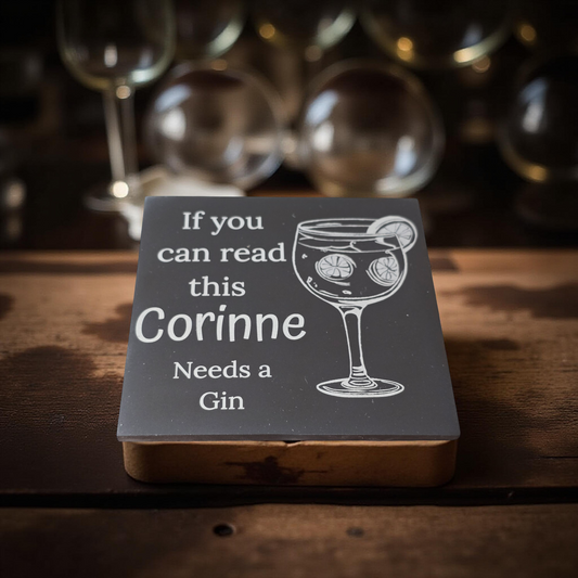 Personalized coaster with a glass of gin and text on a wooden surface with blurred glasses in the background.