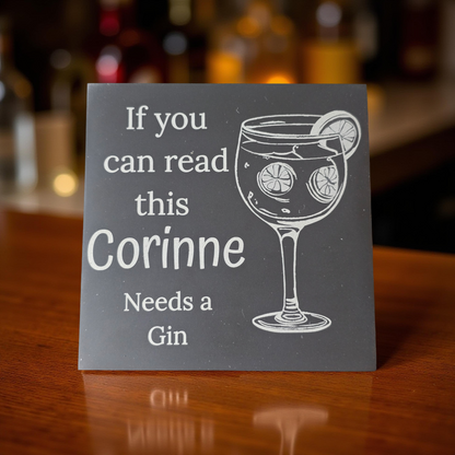 Personalized slate coaster with a glass of gin and text on a wooden surface.