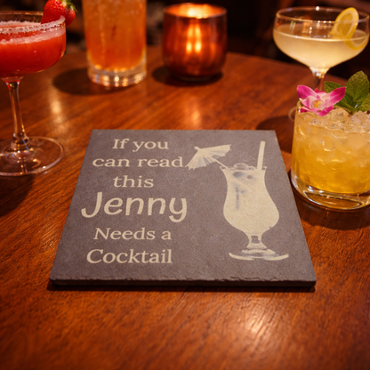 Slate coaster with 'If you can read this Jenny Needs a Cocktail' text and engraving of a cocktail glass on a bar setting with drinks.