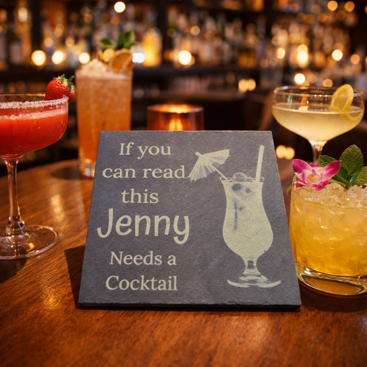 Personalised slate coaster with 'If you can read this Jenny Needs a Cocktail' text on a bar setting with cocktails.