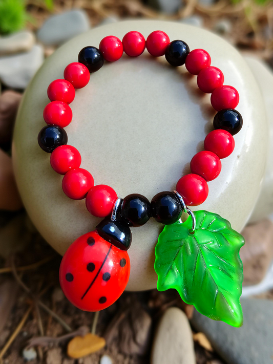 Kids Ladybird Bracelet with red and black beads, a ladybird charm, and a green leaf charm on a stone surface.