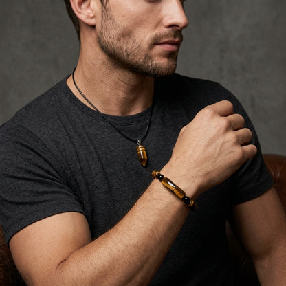 A man wearing a black cord necklace with a brown tigers eye bullet pendant and a matching brown beaded bracelet on his wrist, paired with a dark grey t-shirt against a textured grey background.